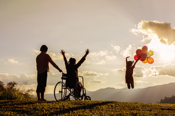 Little girl jumping holding multicolored balloons, mother in wheelchair with raised arms looking at...