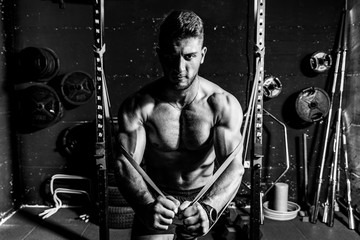 Fototapeta na wymiar Young sweaty strong focused fit muscular man chest stretching cross workout training in improvised gym with rubber for strength and good looking of muscles dark image black and white