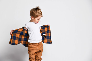 Little kid boy in t-shirt and jeans is holding opened taking off his plaid shirt looking at it down...