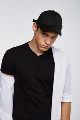 Cropped front view shot of a dark-haired man, wearing black baseball cap, black t-shirt and white shirt. He is posing on grey background and looking straight. 