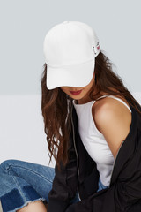 Cropped upward shot of a dark-haired girl, wearing white baseball cap, white tank top, jeans with scuffs and black jacket. The cap peak is closing upper part of a face. 
