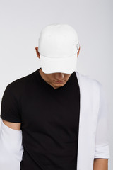 Cropped upward shot of a dark-haired man, wearing white baseball cap, black t-shirt and white shirt. The cap peak is closing upper part of a face. He is posing on grey background. 