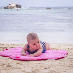 White baby on the beach lying in the sand on pink mat watching and enjoying the sea and blue sky