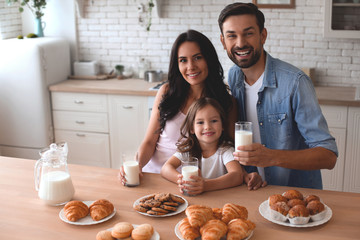 cute girl, mother and father holding cookies with milk glasses and looking at the camera and in the kitchen at home