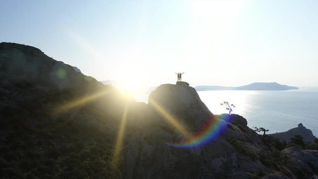 Aerial silhouette of a loving couple standing on the top of a mountain over the sea at sunrise and waving hands. Flight over Young active mountaineers man and woman on the summit in Crimea