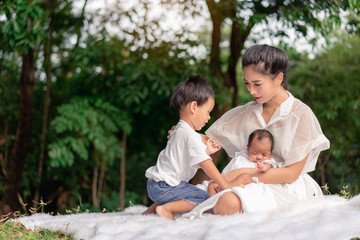 Happy loving family. Asian young beautiful mother and her children, new born baby girl and a boy sitting on green grass to playing and hugging in the park