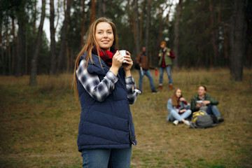 Close up of woman drinking coffee or tea with group of friends on a hiking trip in autumn day. People with touristic bags in the forest, talking, laughting. Leisure activity, friendship, weekend.