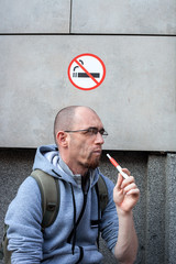 Caucasian man smoke Iqos cigarette under sign No smoking, concept violation of rules