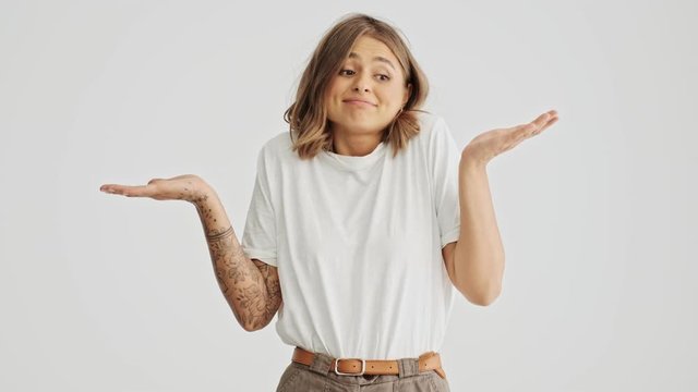 Puzzled and confused young woman wearing a basic t-shirt spreading her hands to the sides while shrugging shoulders isolated over white background