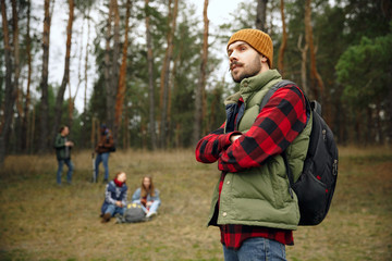 Close up of man hands crossed with background of group of friends on a hiking trip in autumn day. People with touristic bags in the forest, talking, laughting. Leisure activity, friendship, weekend.