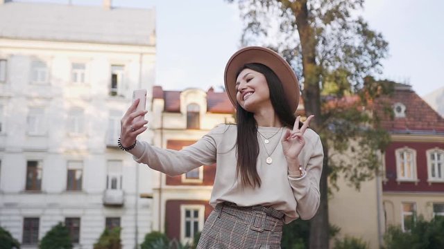Good-looking smiling slender modern 30-aged girl in stylish clothes and hat which posing on her phone and making funny photos on the background of the beautiful rich mansions