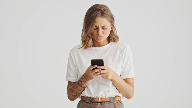 Displeased young woman wearing basic t-shirt using her smartphone isolated over white background