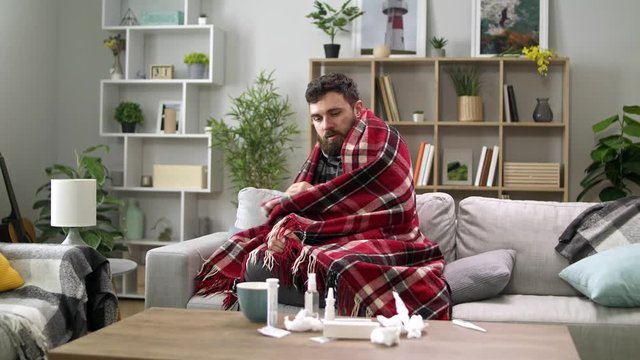 Ill man with chills and wraps himself in a blanket on the sofa in living room