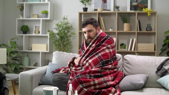 Ill man with chills and wraps himself in a blanket on the sofa in living room