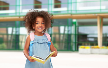 childhood and education concept - happy little african american girl with book and backpack over school yard background