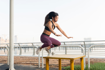 Beautiful brunette fitness model wearing a black sports top and purple tights does a box jump exercise on a bright sunny day.