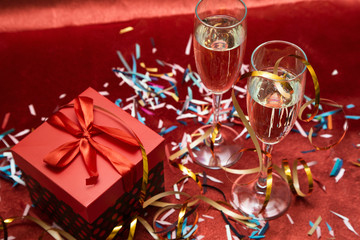 Two glasses with champagne and a gift box stand on a red background with confetti and serpentine. Greeting card for New Year and Christmas. Romantic postcard.