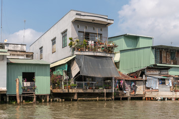 Fototapeta na wymiar Cai Be, Mekong Delta, Vietnam - March 13, 2019: Along Kinh 28 canal. Rather nice green and gray dwellings built half over the brown water under blue cloudscape. 