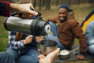 Naklejka premium Group of friends on a camping or hiking trip in autumn day. Men and women with touristic backpacks having break in the forest, talking, laughting. Leisure activity, friendship, weekend. Eat and drink.