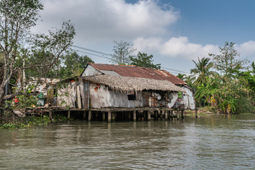 Fototapeta na wymiar Cai Be, Mekong Delta, Vietnam - March 13, 2019: Along Kinh 28 canal. Hut-like with straw roof poor houses built on stilts and on shore with extra corrugated roof plates under blue cloudscape and green