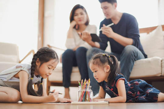 Young Asian family love, daughter sitting and laying on the wood floor in living room, education to writing and painting which smiling and felling happy near father and mother sit on sofa at home