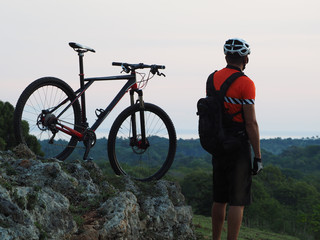 A cyclist stands on top of a hill, with a mountain bike at dusk. On the horizon the Atlantic Ocean. Active lifestyle.