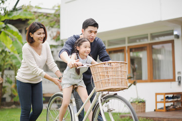 Young Asian family love, father and mother teach and training daughter to ride a bike on grass field at outdoor of home. girl train to ride a bike by her parent which smiling and felling happy