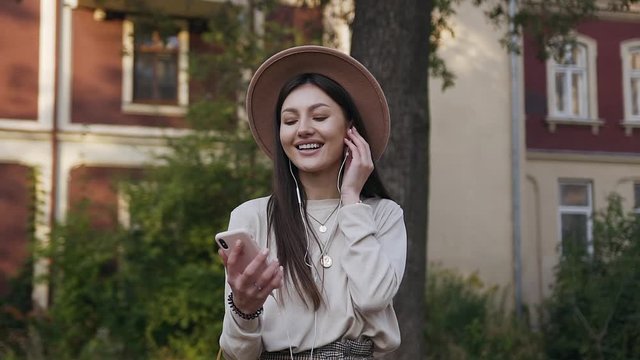 Happy cheerful good-looking woman in beige hat listening nice music on headphones during her walk near beautiful mansion
