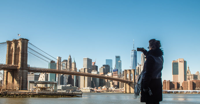 Young Woman tourist by the river in Dumbo taking pictures of Brooklyn Bridge and Cityscape of New York skyline.