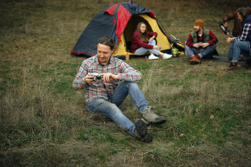 Group of friends on a camping or hiking trip in autumn day. Men and women with touristic bag having break in the forest, talking, laughting. Leisure activity, friendship, weekend. Drink in thermos.