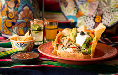 Crispy fajita tortilla tostada bowl with chicken, sautéed bell peppers, onions, tomato, topped with guacamole, sour cream, and olive. Served with chips and dipping salsa styled on a sarape.