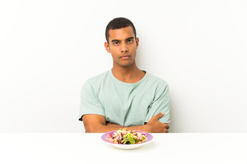 Young handsome man with salad in a table keeping arms crossed