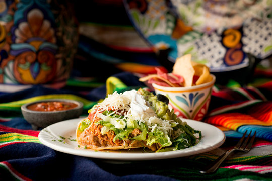 Crispy tortilla tostada with chicken, beans, lettuce, cheese, guacamole, sour cream, and salsa. Served with chips and dipping salsa styled on a sarape. - Powered by Adobe