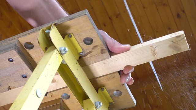 Close POV shot from below of a man’s hands sawing the end off a piece of wood, which is clamped in a portable workbench.