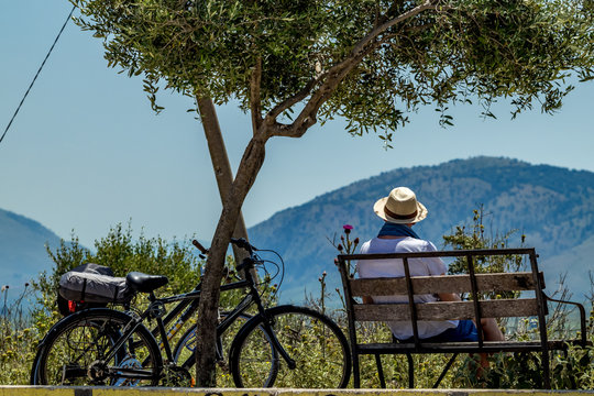 Male fit biker takes some rest and enjoys scenery Albanian views on a bench with his bike under tree shadow. Close to Butrint, Albania, sunny warm spring day