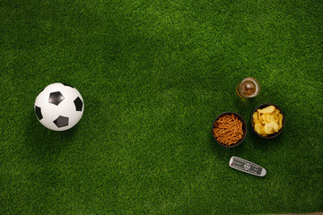 Naklejka premium Soccer ball on a green field and ottoman for a fan with snacks and a TV remote control. flat lay.Copy space.