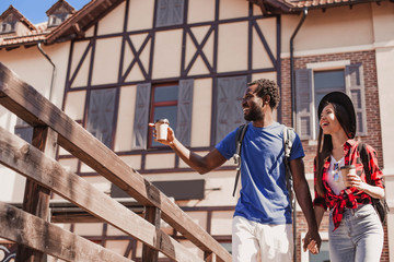 Obraz na płótnie Canvas african american man holding coffee cup and showing something to his girlfriend outdoors
