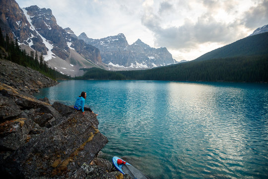A woman takes a break from stand up paddle boarding to take in the views at Moraine Lake in Banff National Park in Canada at dusk in the summer.