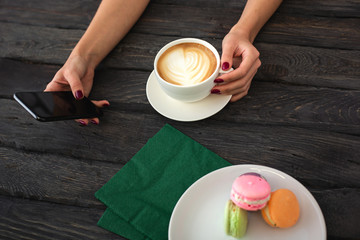 Woman holds phone in one hand and cup of cappuccino in another. Macaroons on .a dark wooden table.