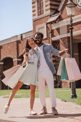 multiracial couple with shopping bags and taking selfie with smartphone using selfiestick on the street