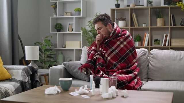 A man coughs heavily while sitting in a plaid on a couch in the living room