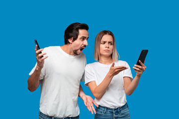 Brunet man in white tees and blue jeans is shocked looking at the smartphone screen of a blond woman