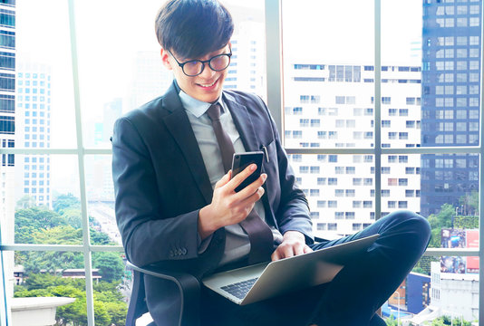 Businessman sitting and holding smartphone on right hand and place laptop on his legs near the window