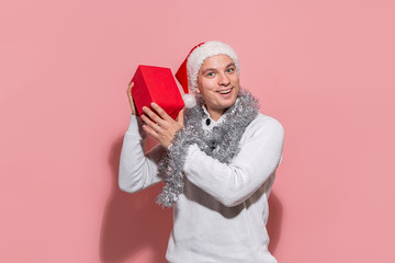 Man in a white sweater and red Santa hat shaking red box to find out what gifts are in.