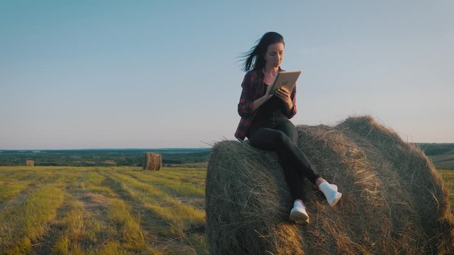 Girl farmer uses tablet in the field next to haystack at sunset. Smart farming, using modern technologies in agriculture.