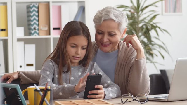 Medium shot of attractive aged Caucasian woman sitting at desktop and using cellphone with the help of her granddaughter sitting nearby. Then they looking at cameraMedium shot of attractive aged Cauca
