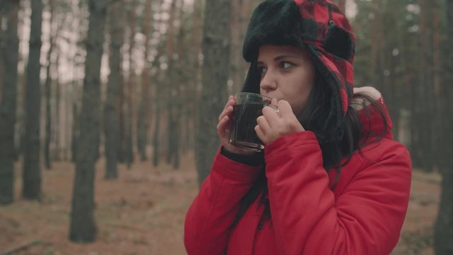 A portrait of the pretty woman with a cup of tea in woods. The woman in a jacket and a hat with earflaps is drinking a tea in a cold weather.