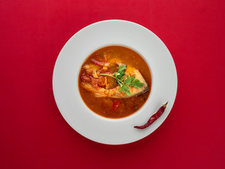 Fish soup in white plate with chilli pepper on red background. Overhead view.