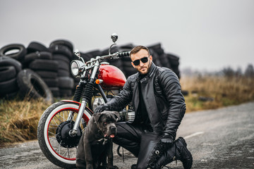 Fototapeta na wymiar Biker in a leather suit crouched near his dog and red motorcycle on the road. Many tires on the background