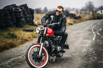 Fototapeta na wymiar Bearded man in sunglasses and leather jacket smiling while sitting on a red motorcycle on the road. Behind him is a row of tires